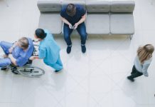 High Angle Shot in the Hospital Lobby, Young Man Waits for Results while Sitting and Using Mobile Phone, Doctors, Nurses and Patients Walk Past Him. Clean, New Hospital.