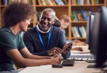 Male University Or College Student Working At Computer In Library Being Helped By Tutor