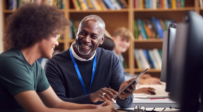 Male University Or College Student Working At Computer In Library Being Helped By Tutor