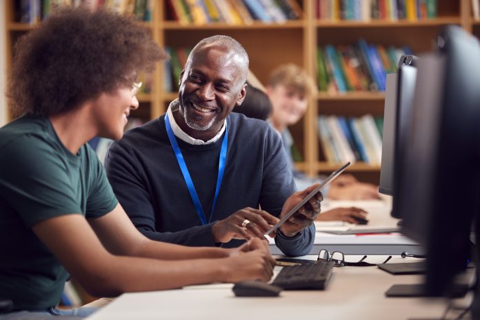 Male University Or College Student Working At Computer In Library Being Helped By Tutor