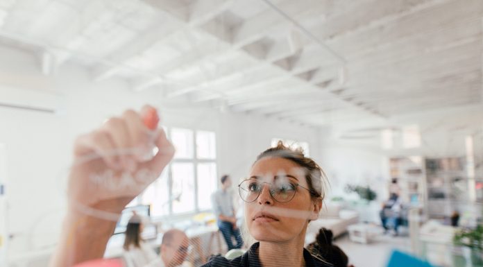 Photo of a young woman writing on a transparent wipe board and thinking of a solution for her work-related problems