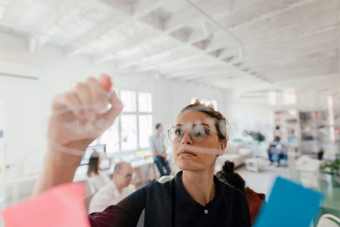 Photo of a young woman writing on a transparent wipe board and thinking of a solution for her work-related problems