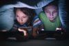 Boy and girl playing games on mobile phones while lying on bed in bedroom under the blanket. They are spending some nice time together that makes them happy.