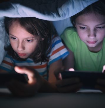 Boy and girl playing games on mobile phones while lying on bed in bedroom under the blanket. They are spending some nice time together that makes them happy.