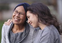 A senior woman with cancer is embraced and comforted by her adult daughter as they sit outside on a fall evening. The mother is smiling and laughing while the daughter is squeezing her mother affectionately and smiling as well.