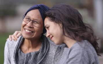 A senior woman with cancer is embraced and comforted by her adult daughter as they sit outside on a fall evening. The mother is smiling and laughing while the daughter is squeezing her mother affectionately and smiling as well.