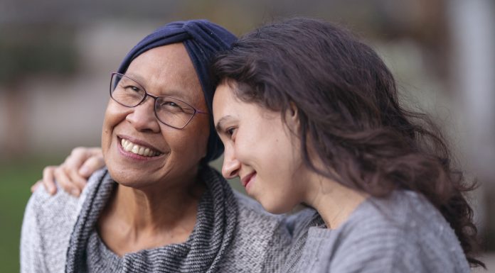 A senior woman with cancer is embraced and comforted by her adult daughter as they sit outside on a fall evening. The mother is smiling and laughing while the daughter is squeezing her mother affectionately and smiling as well.