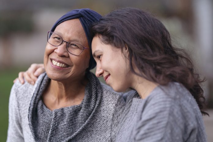 A senior woman with cancer is embraced and comforted by her adult daughter as they sit outside on a fall evening. The mother is smiling and laughing while the daughter is squeezing her mother affectionately and smiling as well.