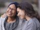 A senior woman with cancer is embraced and comforted by her adult daughter as they sit outside on a fall evening. The mother is smiling and laughing while the daughter is squeezing her mother affectionately and smiling as well.