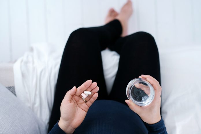 Personal perspective of pregnant woman sitting on bed side at home, taking medicines in hand with a glass of water. Essential vitamins and supplements are vital for the healthy growth and development of the unborn baby