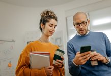 Mid adult bald businessman with coffee cup showing smart phone to smiling female coworker while standing in office