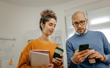 Mid adult bald businessman with coffee cup showing smart phone to smiling female coworker while standing in office