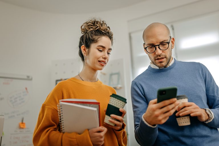 Mid adult bald businessman with coffee cup showing smart phone to smiling female coworker while standing in office