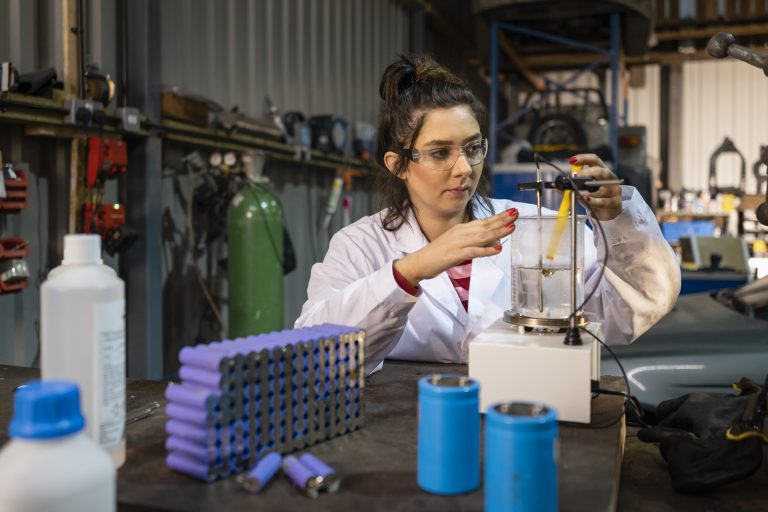 A medium close up view of a female mechanic working in a garage and recycling lithium batteries she is using hazardous materials and is using laboratory glassware for her work. She is also using an electric hotplate stirrer in the process of recycling.