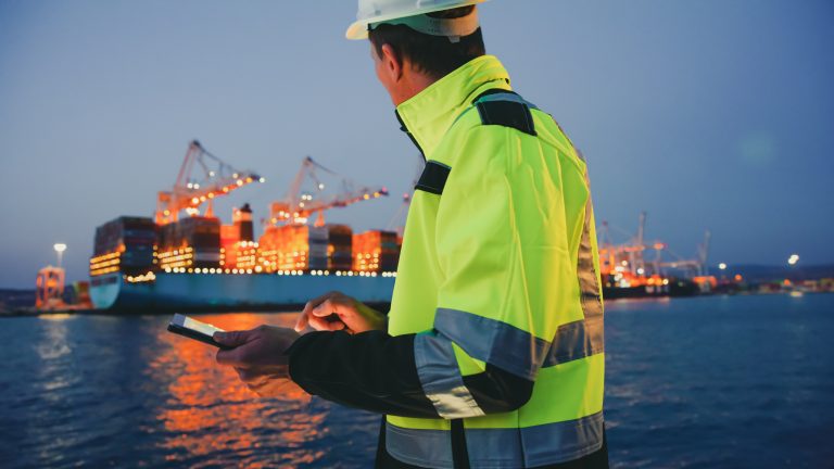 Man in protective gear holding tablet computer in front of harbor terminal at night