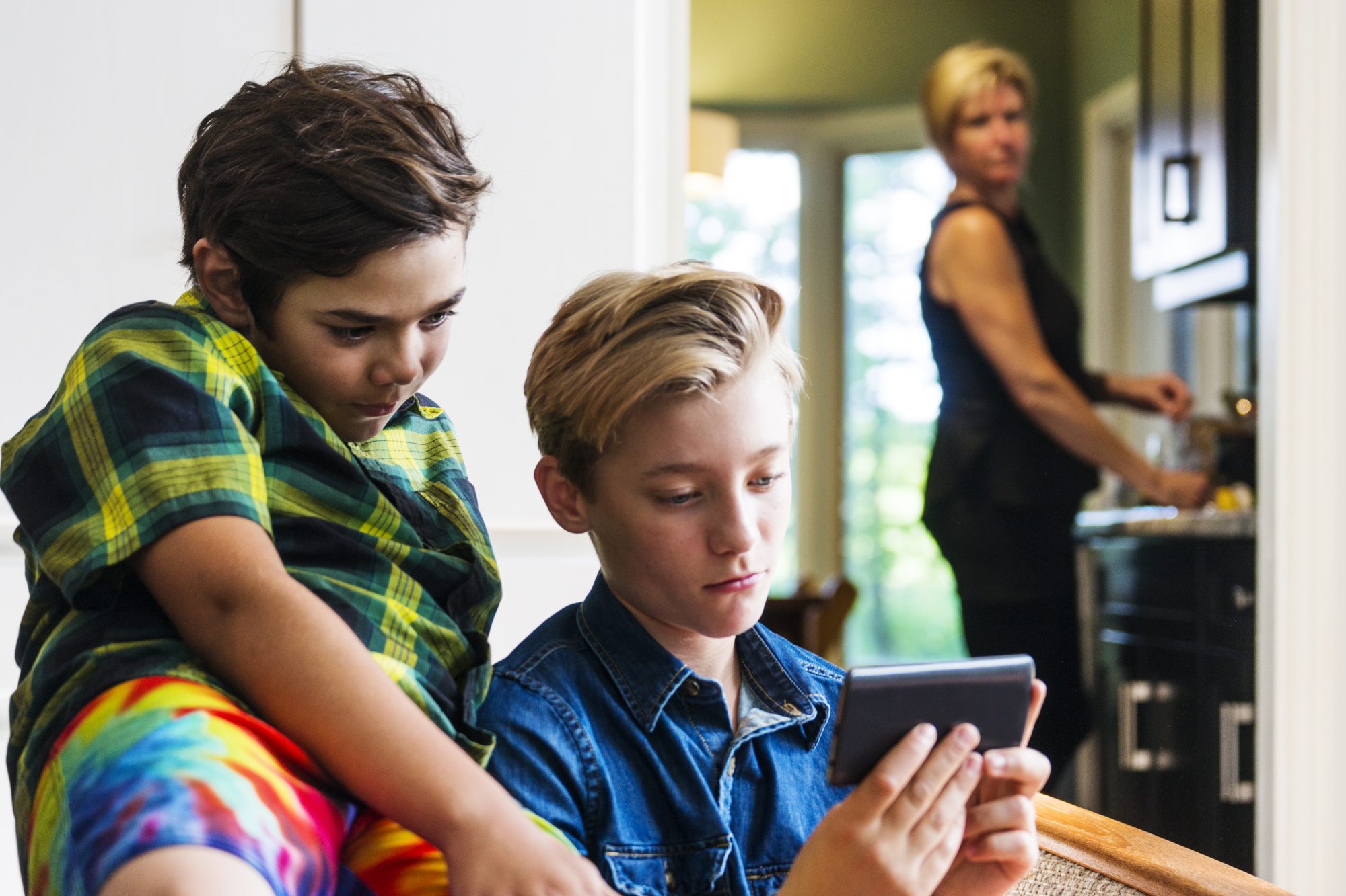 A mother looks on from the kitchen as two pre-teen boys use a smartphone in the living room. She is out of focus but it is clear to see her sense of discomfort with what they are doing on the internet.