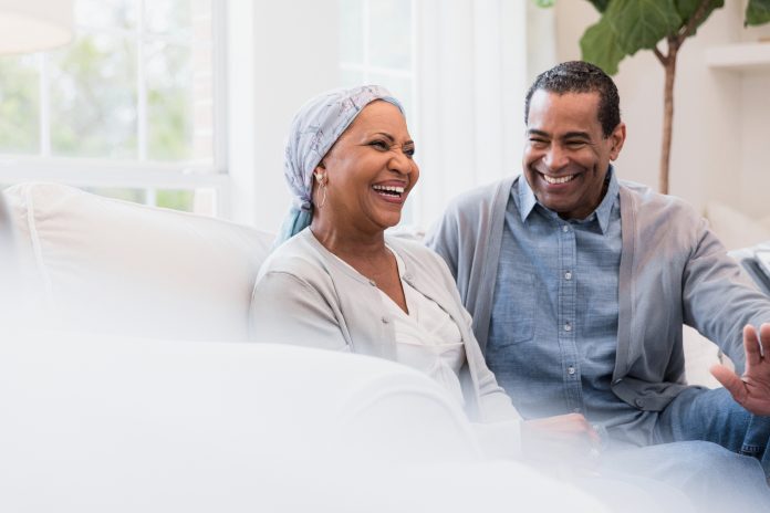 Sitting on their sofa at home, the senior adult couple smiles and laughs at a joke.