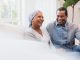 Sitting on their sofa at home, the senior adult couple smiles and laughs at a joke.