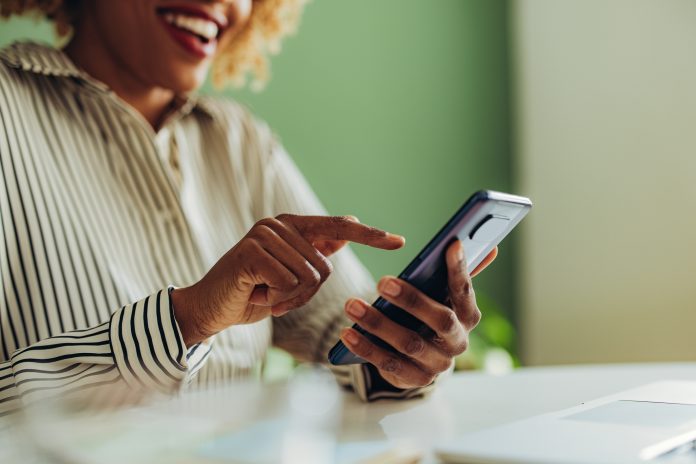 An anonymous African American woman typing text message on her smartphone while sitting at office desk with laptop computer.