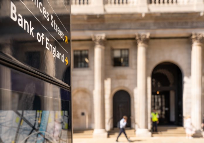 Close-up on a sign for the Bank of England, with a pedestrian passing the institution's main entrance in the background.