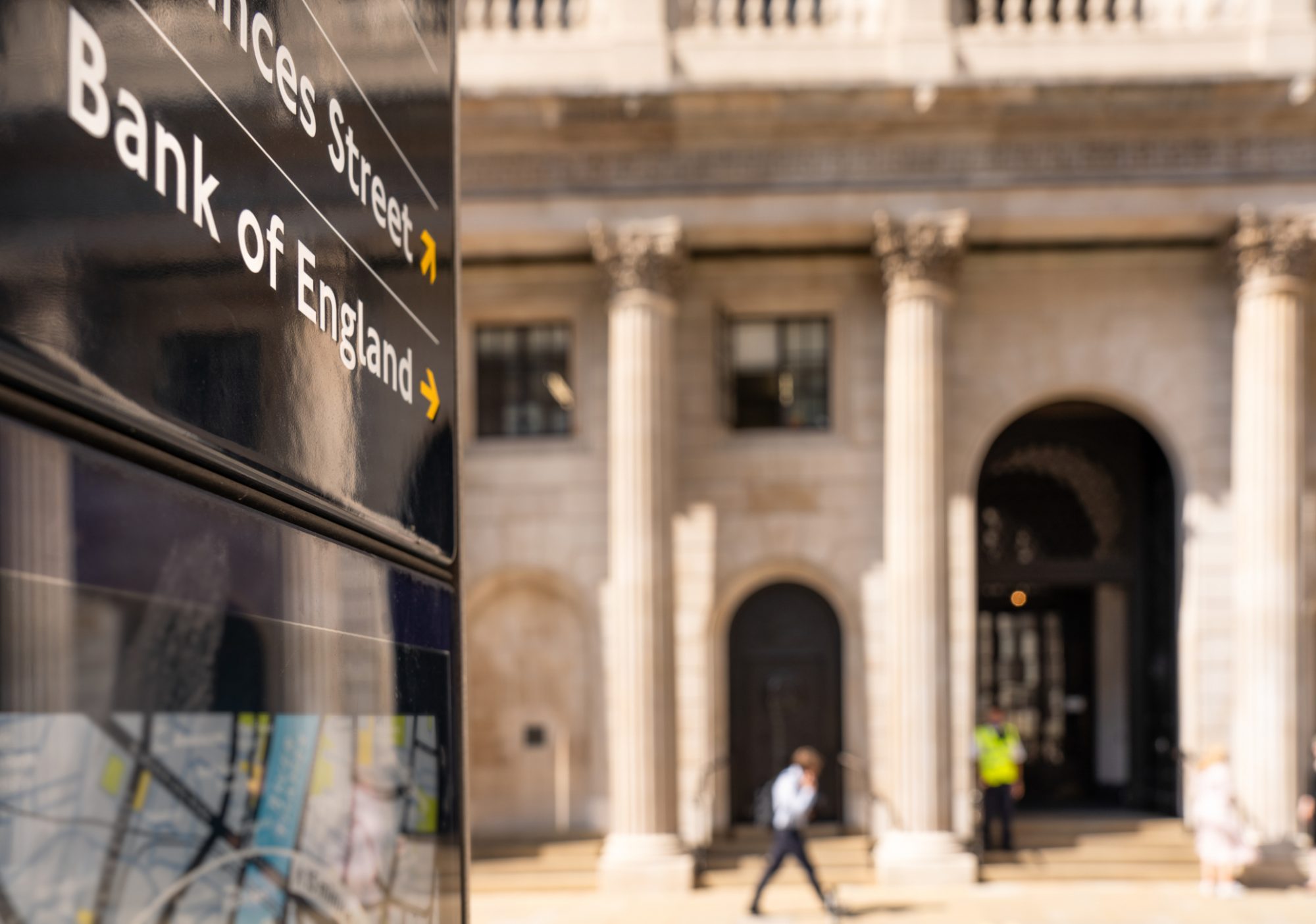 Close-up on a sign for the Bank of England, with a pedestrian passing the institution's main entrance in the background.
