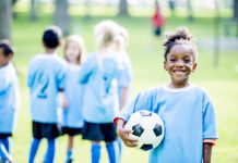 A group of kids wearing soccer uniforms are outdoors on a summer day. A girl of African descent in the foreground is smiling at the camera while holding, community sport