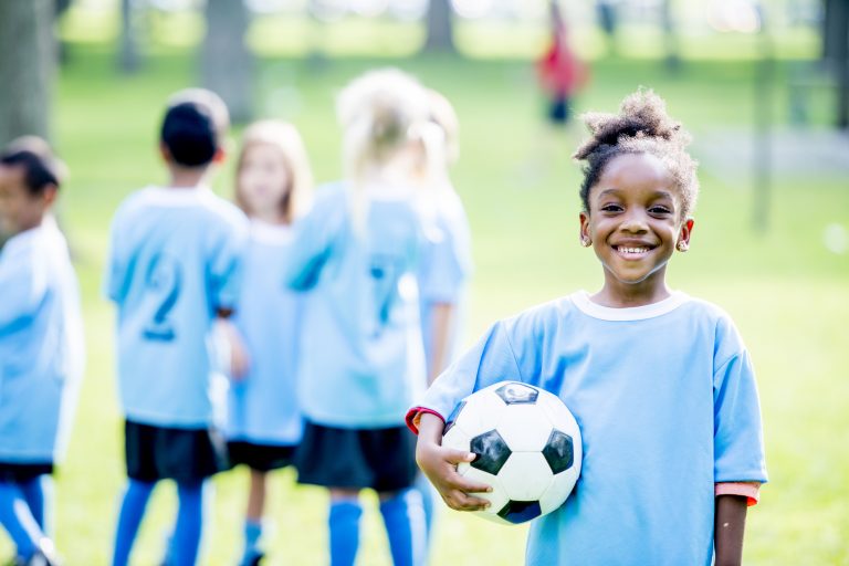 A group of kids wearing soccer uniforms are outdoors on a summer day. A girl of African descent in the foreground is smiling at the camera while holding, community sport
