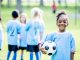 A group of kids wearing soccer uniforms are outdoors on a summer day. A girl of African descent in the foreground is smiling at the camera while holding, community sport