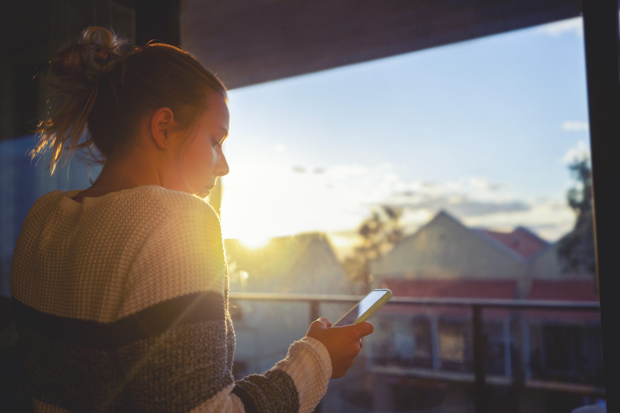 Woman looking through the window at sunset. She is alone and looks a little sad or depressed. She is using her mobile phone.