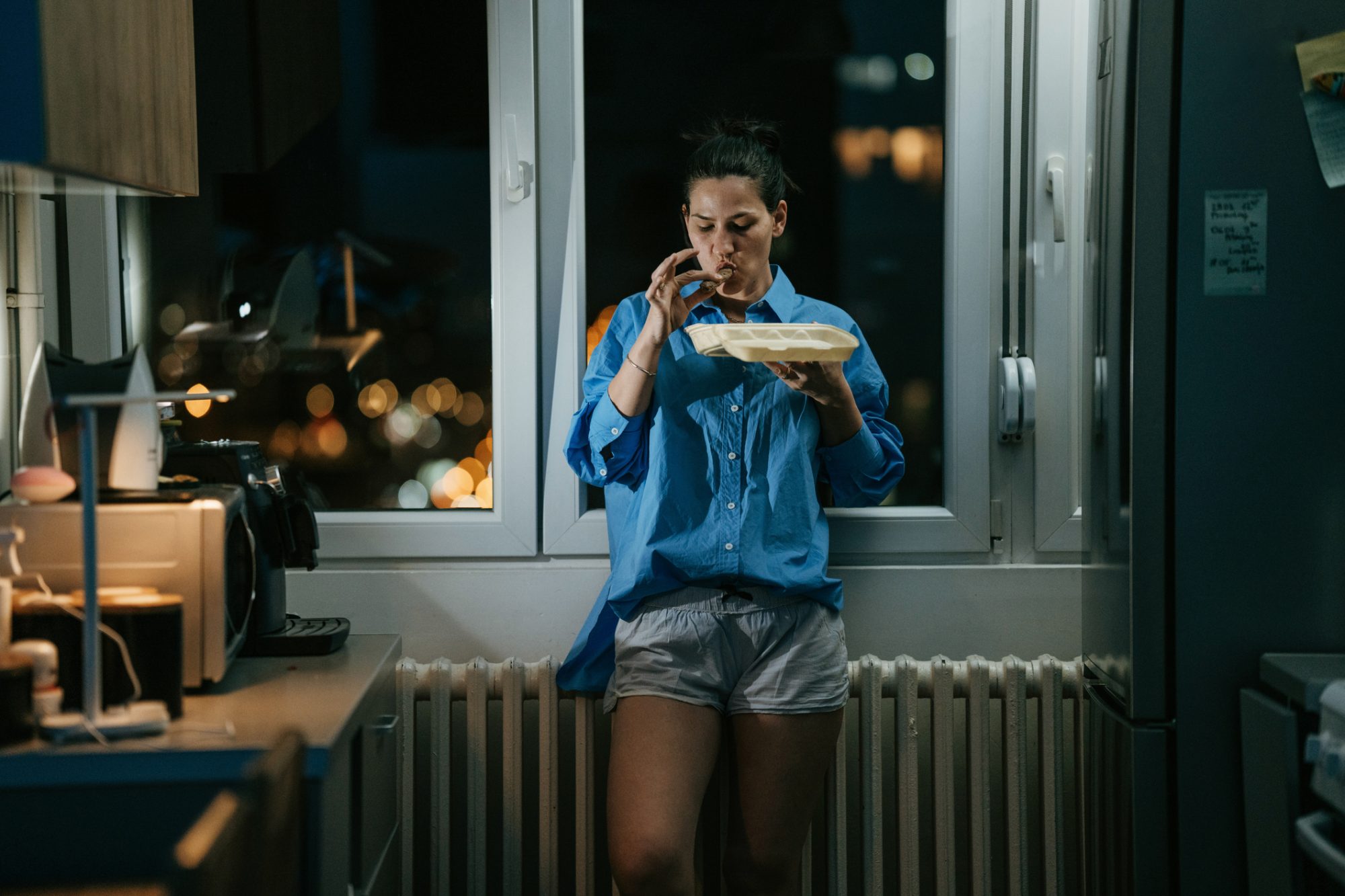 Woman standing in kitchen late at night, she is eating take out food alone.