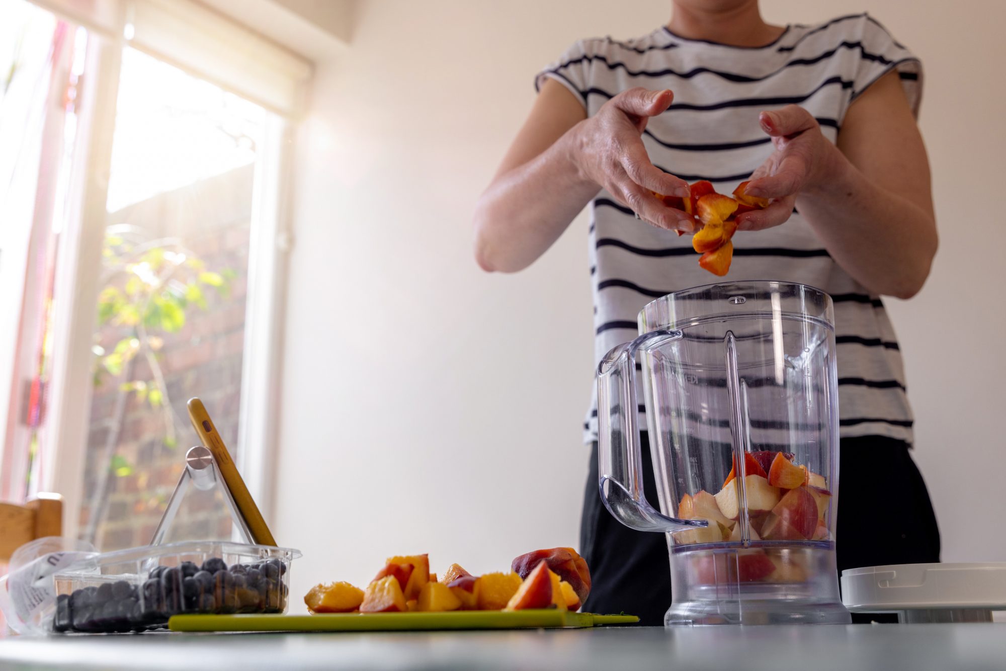 Medium shot, part of a mature female adult putting fruit into a fruit blender in her kitchen with the sun coming in through the window. She has her phone stood up reading a recipe from it.