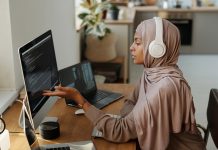 Young Muslim female programmer pointing at data on computer screen while sitting by workplace and communicating with colleague in video chat, flexible work