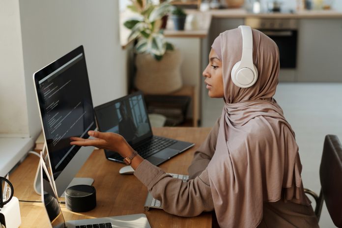 Young Muslim female programmer pointing at data on computer screen while sitting by workplace and communicating with colleague in video chat, flexible work