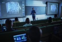 Young University Professor Explaining the Importance of Artificial Intelligence to a Group of Diverse Multiethnic Students in a Dark Auditorium. Teacher Showing Neural Network on Two Big Screens, cyber skills and learning