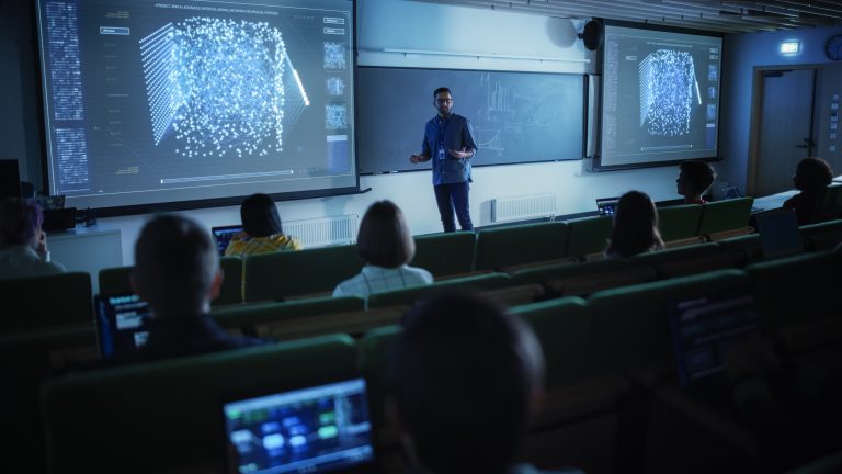 Young University Professor Explaining the Importance of Artificial Intelligence to a Group of Diverse Multiethnic Students in a Dark Auditorium. Teacher Showing Neural Network on Two Big Screens, cyber skills and learning