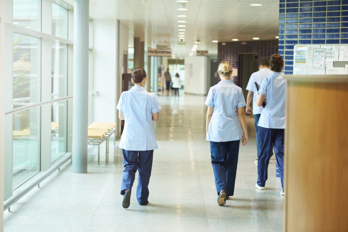 a group of four young trainee nurses including male and female nurses , walk away from camera down a hospital corridor . They are wearing uk nurse uniforms of trousers and tunics.