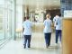 a group of four young trainee nurses including male and female nurses , walk away from camera down a hospital corridor . They are wearing uk nurse uniforms of trousers and tunics.