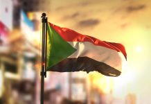 Sudan Flag Against City Blurred Background At Sunrise Backlight