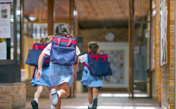 Rear view of excited students running towards entrance. Girls are carrying backpacks while leaving from school. Happy friends are wearing school uniforms, childhood education