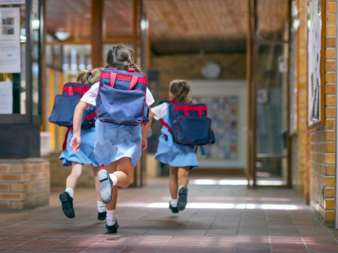 Rear view of excited students running towards entrance. Girls are carrying backpacks while leaving from school. Happy friends are wearing school uniforms, childhood education