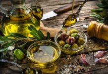 Close up view of two glass bowls filled with olives and extra virgin olive oil shot on rustic wooden table. A spoon pouring oil comes from the top right. Olive tree branches, garlic, salt and peppercorns complete the composition. High resolution 42Mp studio digital capture taken with Sony A7rII and Sony FE 90mm f2.8 macro G OSS lens