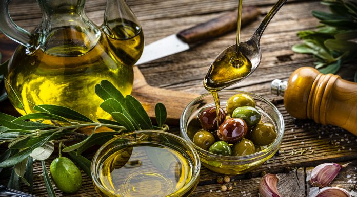 Close up view of two glass bowls filled with olives and extra virgin olive oil shot on rustic wooden table. A spoon pouring oil comes from the top right. Olive tree branches, garlic, salt and peppercorns complete the composition. High resolution 42Mp studio digital capture taken with Sony A7rII and Sony FE 90mm f2.8 macro G OSS lens