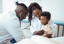 A happy toddler boy sits in his mother' lap at the pediatrician. He plays patacake with a doctor. Doctor giving prescription to african american mother with baby son at clinic. Medicine, healthcare and pediatry concept.