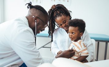 A happy toddler boy sits in his mother' lap at the pediatrician. He plays patacake with a doctor. Doctor giving prescription to african american mother with baby son at clinic. Medicine, healthcare and pediatry concept.