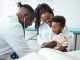 A happy toddler boy sits in his mother' lap at the pediatrician. He plays patacake with a doctor. Doctor giving prescription to african american mother with baby son at clinic. Medicine, healthcare and pediatry concept.