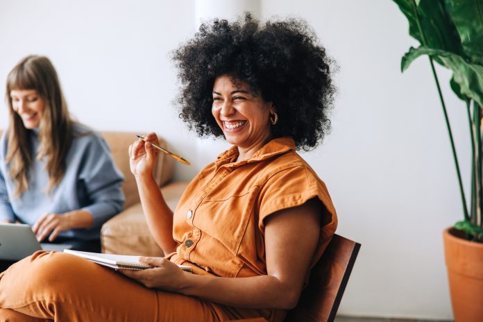 Black businesswoman smiling cheerfully while sitting in a meeting with her colleague. Happy young businesswomen working together in a modern workplace - female entrepreneurship