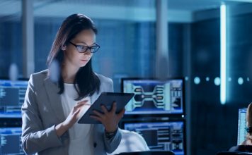 Young Female Government Employee Wearing Glasses Uses Tablet in System Control Center. In the Background Her Coworkers are at Their Workspaces with many Displays Showing Valuable Data.