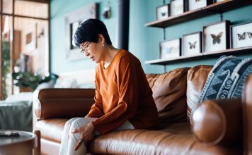 A distraught senior Asian woman feeling unwell, suffering from pain in leg while sitting on sofa in the living room at home