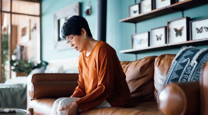 A distraught senior Asian woman feeling unwell, suffering from pain in leg while sitting on sofa in the living room at home