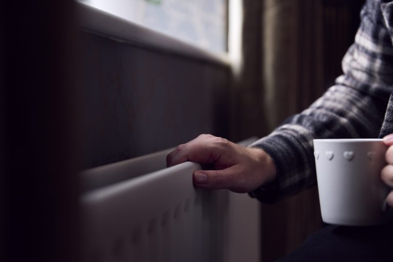 Close Up Of Mature Man Trying To Keep Warm By Radiator At Home During Cost Of Living Energy Crisis